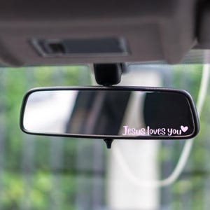 May include: A black car rearview mirror with the text "Jesus loves you" in a pink script font, accompanied by a small pink heart. The mirror is mounted inside a vehicle.