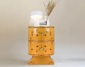 May include: A two-drawer nightstand with a rounded top and a base. The nightstand is made of wood with a light brown finish and has a decorative pattern. The nightstand has silver knobs on the drawers. A white lamp and a digital clock are on top of the nightstand.
