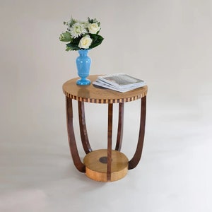 May include: A round wooden side table with a light wood top and dark wood legs. The table has a lower shelf and is decorated with a geometric pattern. A blue vase with white flowers and a stack of magazines are on the table.
