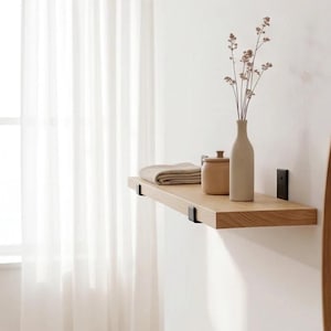 May include: A light-toned wooden wall shelf mounted with black metal brackets. The shelf holds a folded beige cloth, a small tan lidded container, and a tall, slender beige vase containing a delicate, dried botanical stem with tiny flowers.