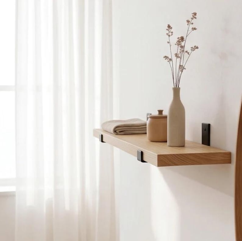 May include: A light-toned wooden wall shelf mounted with black metal brackets. The shelf holds a folded beige cloth, a small tan lidded container, and a tall, slender beige vase containing a delicate, dried botanical stem with tiny flowers.