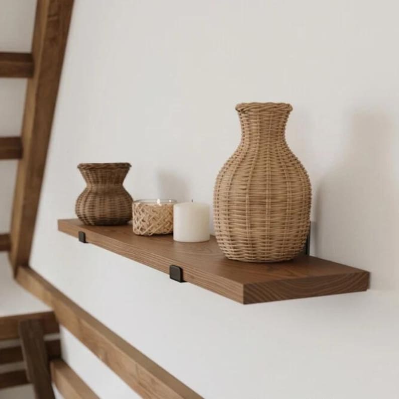 May include: A horizontal wooden shelf mounted on a white wall, holding two woven rattan vases of different sizes, a small candle in a woven holder, and a plain white cylindrical candle. The shelf is secured with black metal brackets.