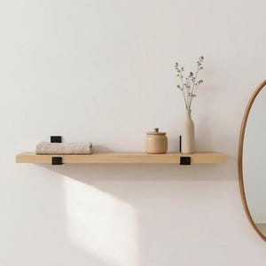 May include: A light-toned wooden wall shelf mounted with two black metal brackets. On the shelf sits a folded beige textile, a small tan ceramic jar with a lid, and a tall, slender beige vase containing a sprig of dried, delicate flowers.