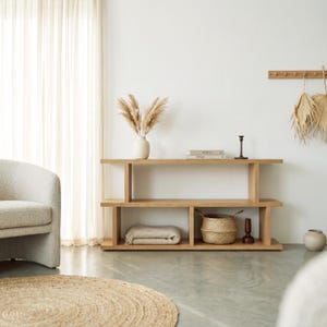 May include: A light-toned wooden console table with two horizontal shelves. The top shelf holds a white vase with dried pampas grass, two stacked books, and a black candle holder. The bottom shelf contains a folded beige blanket and a woven basket.