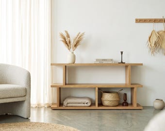 May include: A light-toned wooden console table with two horizontal shelves. The top shelf holds a white vase with dried pampas grass, two stacked books, and a black candle holder. The bottom shelf contains a folded beige blanket and a woven basket.