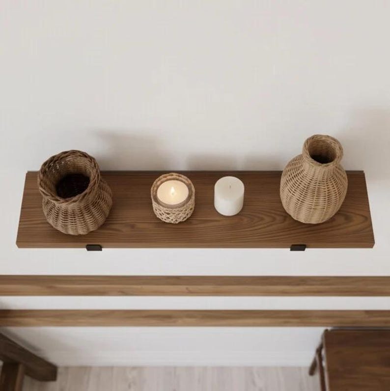 May include: A horizontal wooden wall shelf with two black metal brackets holding a woven wicker basket, a candle in a woven holder, a plain white pillar candle, and a woven wicker vase. The shelf features a visible wood grain pattern.