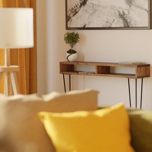 May include: A wooden console table with a shelf and black hairpin legs. The table is decorated with a potted plant and a book. The table is in front of a sofa with yellow and beige cushions.