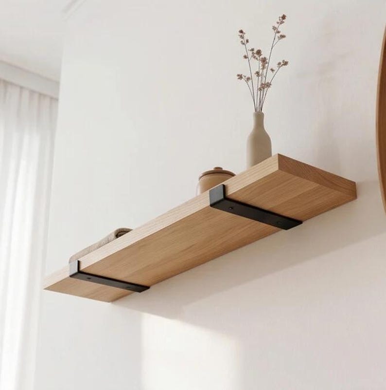 May include: A light-toned wooden wall shelf featuring two matte black metal brackets. The shelf holds a beige ceramic vase containing dried stems and a small, rounded brown container, providing a functional surface for display and storage.