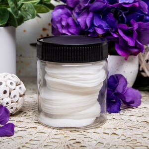 May include: A clear, cylindrical jar with a black lid filled with white cotton pads. The jar is centered on a lace doily, with purple flowers and greenery in the background. The overall aesthetic is clean and spa-like.