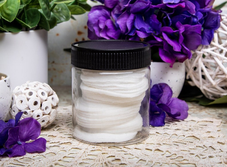May include: A clear, cylindrical jar with a black lid filled with white cotton pads. The jar is centered on a lace doily, with purple flowers and greenery in the background. The overall aesthetic is clean and spa-like.