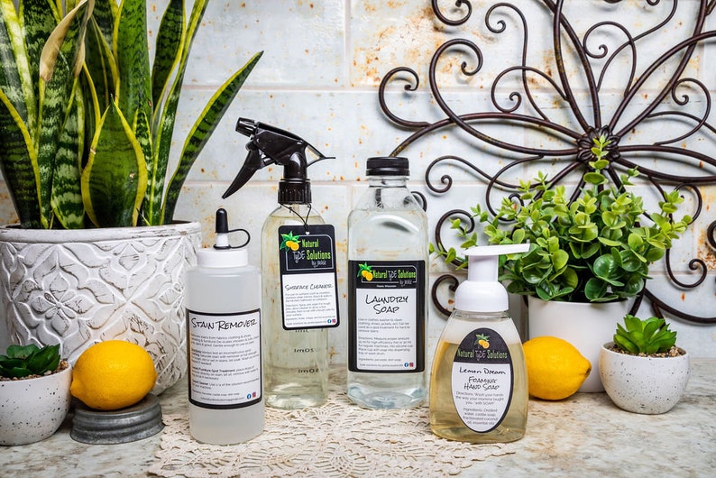 May include: A collection of cleaning products, including stain remover, grease cleaner, laundry soap, and foaming hand soap. The products are in clear bottles with black labels and spray nozzles. The image also includes lemons and potted plants.