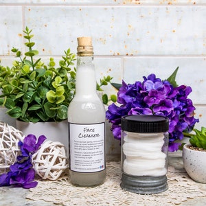May include: A clear glass bottle with a cork stopper and a label that reads "Face Cleanser" sits next to a jar filled with cotton pads. The bottle contains a milky liquid. Decorative purple flowers and greenery surround the products.