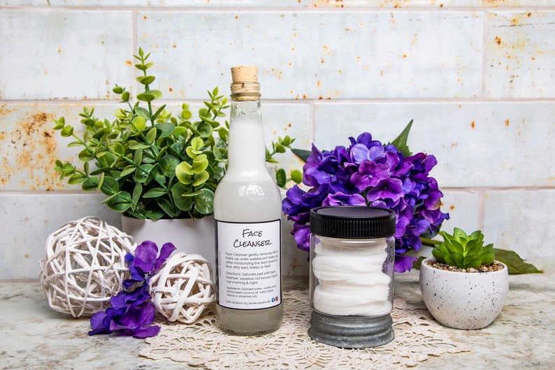 May include: A clear glass bottle with a cork stopper and a label that reads "Face Cleanser" sits next to a jar filled with cotton pads. The bottle contains a milky liquid. Decorative purple flowers and greenery surround the products.