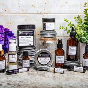 May include: A collection of skincare products, including face cleanser, serums, oils, and lip balm, arranged on a countertop. The products are in amber glass bottles and jars with white labels. Purple flowers and greenery add a decorative touch.