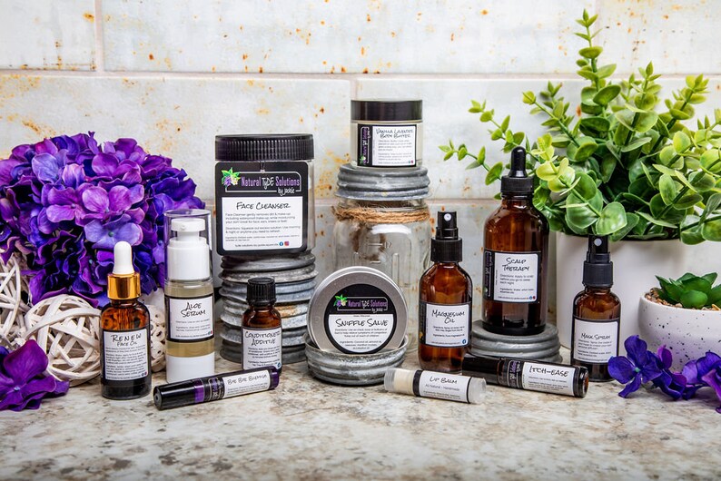 May include: A collection of skincare products, including face cleanser, serums, oils, and lip balm, arranged on a countertop. The products are in amber glass bottles and jars with white labels. Purple flowers and greenery add a decorative touch.