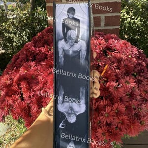 Puede incluir: Un libro negro con una ilustraci&oacute;n en escala de grises de dos figuras en el lomo. El texto "Bellatrix Books" se repite varias veces. El libro se sostiene frente a un fondo de flores rojas.