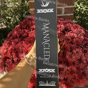 Puede incluir: Un letrero rectangular negro con texto blanco, incluyendo "MANACLED" y "Bellatrix Books". El letrero se sostiene frente a un gran arreglo de flores rojas.
