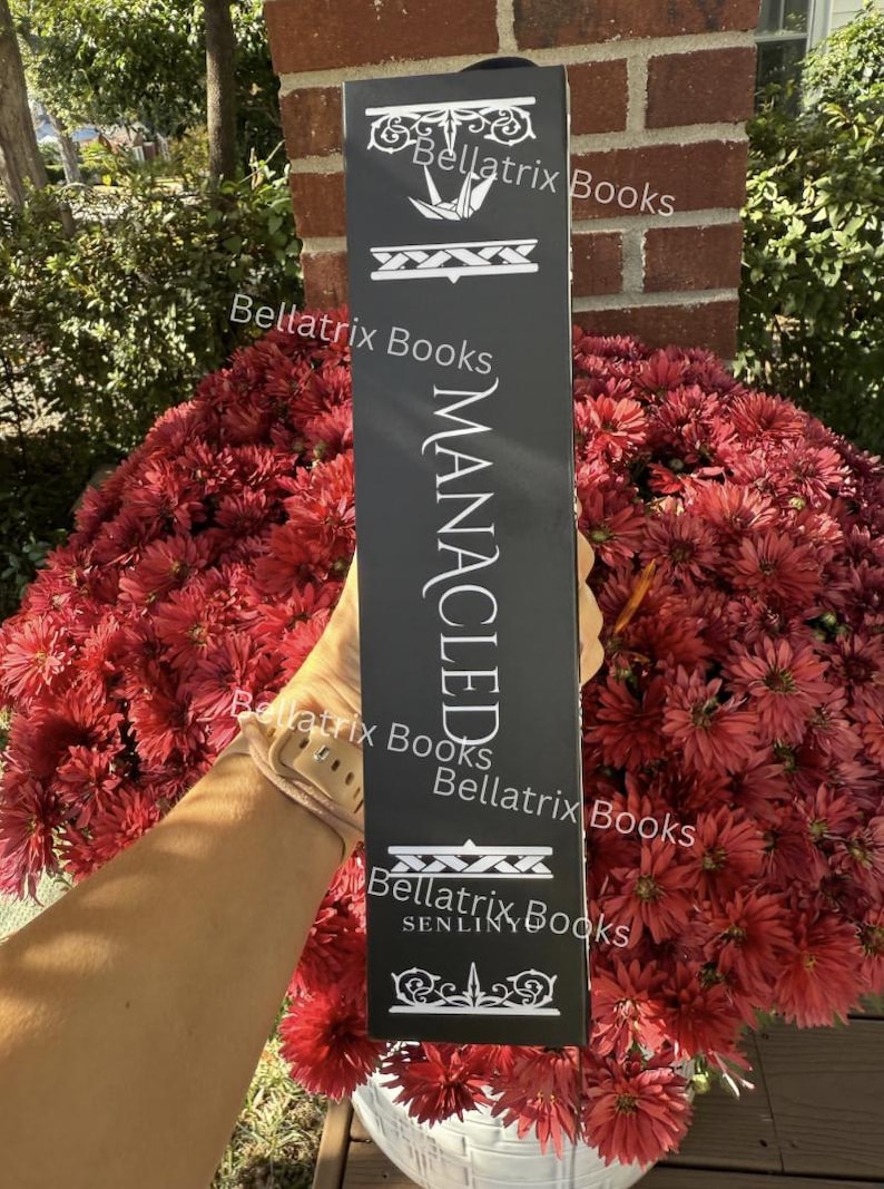 Puede incluir: Un letrero rectangular negro con texto blanco, incluyendo "MANACLED" y "Bellatrix Books". El letrero se sostiene frente a un gran arreglo de flores rojas.