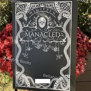 Puede incluir: Cubierta de libro negra con dise&ntilde;o blanco ornamentado con serpientes, una calavera y la palabra "MANACLED". La cubierta tambi&eacute;n incluye el texto "Bellatrix Books" y grullas de origami colgantes. El libro se sostiene frente a un fondo de flores rojas.