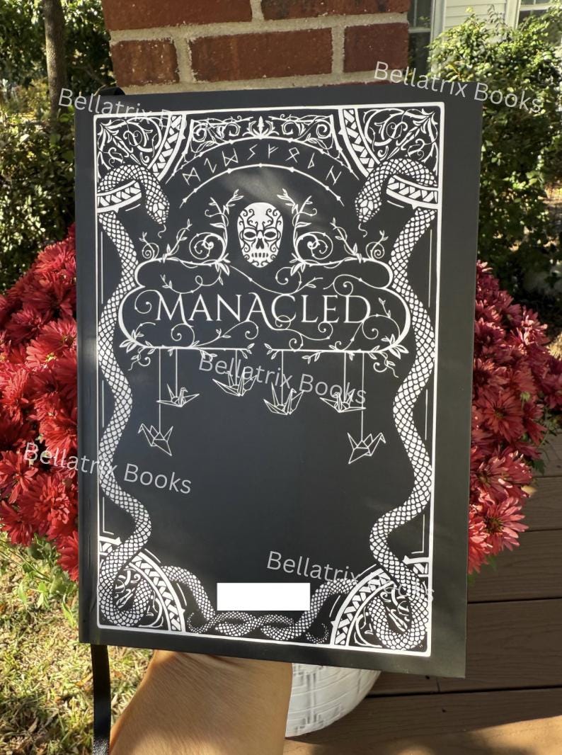Puede incluir: Cubierta de libro negra con dise&ntilde;o blanco ornamentado con serpientes, una calavera y la palabra "MANACLED". La cubierta tambi&eacute;n incluye el texto "Bellatrix Books" y grullas de origami colgantes. El libro se sostiene frente a un fondo de flores rojas.