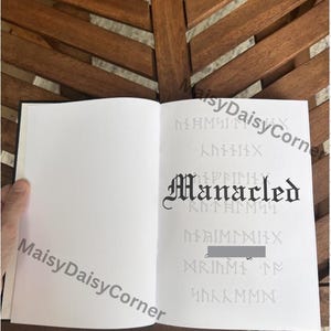 Puede incluir: Un libro abierto con p&aacute;ginas blancas en blanco. La p&aacute;gina derecha presenta la palabra "Manacled" en una fuente g&oacute;tica, con texto y s&iacute;mbolos tenues en el fondo. El libro est&aacute; sobre una superficie de madera.