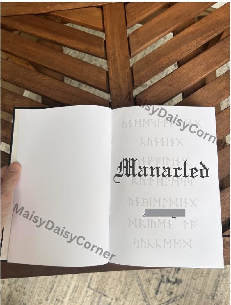 Puede incluir: Un libro abierto con p&aacute;ginas blancas en blanco. La p&aacute;gina derecha presenta la palabra "Manacled" en una fuente g&oacute;tica, con texto y s&iacute;mbolos tenues en el fondo. El libro est&aacute; sobre una superficie de madera.