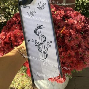 Puede incluir: Un objeto rectangular con marco negro y fondo blanco, con una serpiente negra enrollada alrededor de un bast&oacute;n, detalles florales y grullas de origami. El texto "Bellatrix Books" se repite varias veces. El objeto se sostiene frente a un ramo de flores rojas.