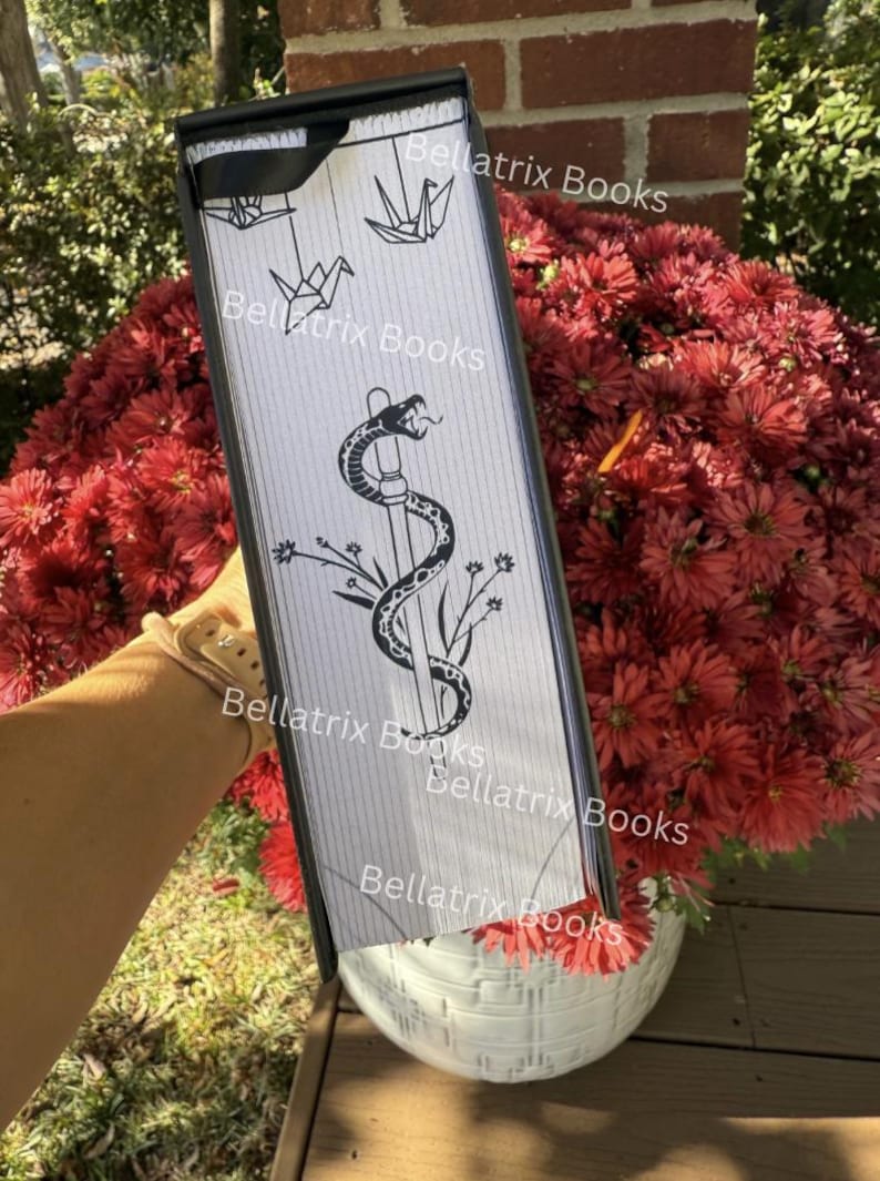 Puede incluir: Un objeto rectangular con marco negro y fondo blanco, con una serpiente negra enrollada alrededor de un bast&oacute;n, detalles florales y grullas de origami. El texto "Bellatrix Books" se repite varias veces. El objeto se sostiene frente a un ramo de flores rojas.