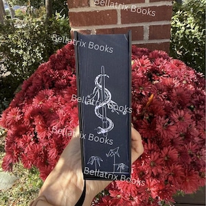 Puede incluir: Un libro negro con el t&iacute;tulo "Bellatrix Books" y una ilustraci&oacute;n blanca de una serpiente enrollada alrededor de una espada. El libro se muestra frente a un gran arreglo de flores rojas. El lomo del libro tambi&eacute;n presenta el t&iacute;tulo.