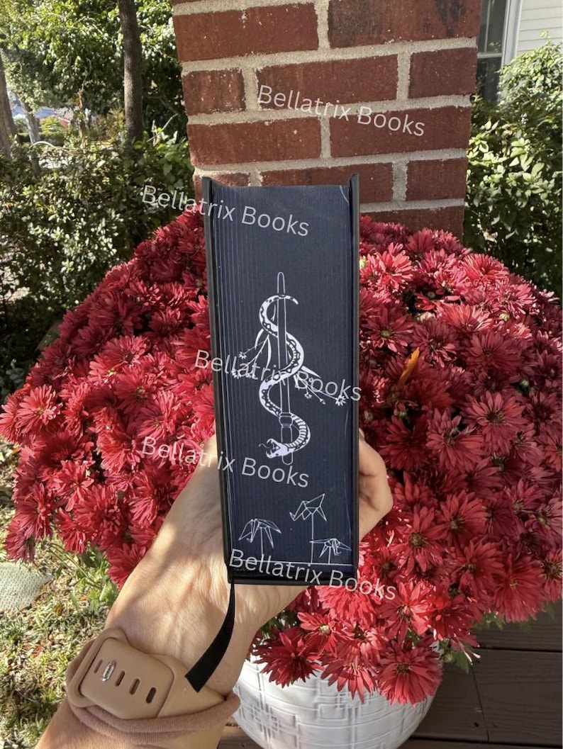 Puede incluir: Un libro negro con el t&iacute;tulo "Bellatrix Books" y una ilustraci&oacute;n blanca de una serpiente enrollada alrededor de una espada. El libro se muestra frente a un gran arreglo de flores rojas. El lomo del libro tambi&eacute;n presenta el t&iacute;tulo.