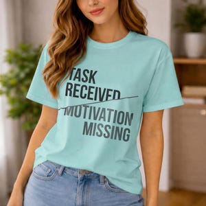 Pode incluir: Camiseta azul clara com o texto "TASK RECEIVED" riscado e "MOTIVATION MISSING" em preto. A camiseta é feita de um material macio e é adequada para uso casual.