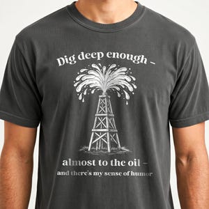 Op de afbeelding: Donkergrijs t-shirt met de tekst "Dig deep enough - almost to the oil - and there's my sense of humor." Een witte illustratie van een boortoren met een uitbarstende geiser staat afgebeeld.