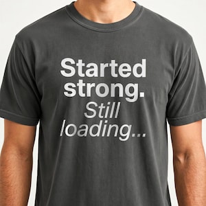 May include: Dark gray t-shirt with the text "Started strong. Still loading..." in white. The shirt has a crew neck and short sleeves. The text is centered on the front of the shirt.