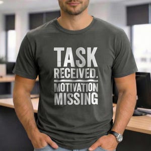 Pode incluir: Camiseta cinza escuro com o texto branco "TASK RECEIVED. MOTIVATION MISSING". A camiseta é de gola redonda e está sendo usada.
