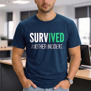 May include: Navy blue crew neck t-shirt with the text "SURVIVED" in white, the "VIVE" in green, and "ANOTHER INCIDENT." in white. The shirt is made of a soft material.