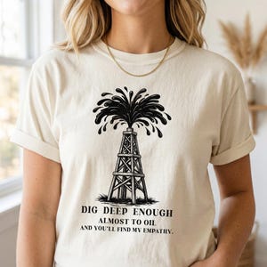 Op de afbeelding: Crèmekleurig T-shirt met een zwarte illustratie van een olieboorinstallatie met olie die eruit spuit. De tekst "DIG DEEP ENOUGH ALMOST TO OIL AND YOU'LL FIND MY EMPATHY" staat eronder.