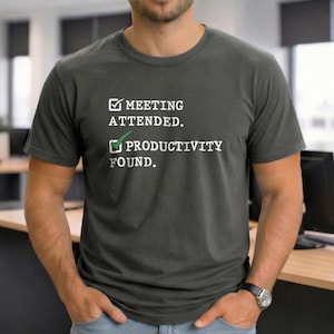 Pode incluir: T-shirt cinza escuro com o texto branco "MEETING ATTENDED." e "PRODUCTIVITY FOUND.". A palavra "PRODUCTIVITY" tem uma marca de seleção verde. A camisa é de gola redonda e mangas curtas.