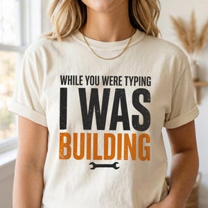 Pode incluir: Camiseta creme com o texto "WHILE YOU WERE TYPING I WAS BUILDING" em preto e laranja. Um gráfico de chave inglesa está abaixo da palavra "BUILDING". A camiseta tem mangas curtas e gola redonda.