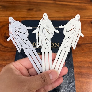 Puede incluir: Tres marcapáginas de papel blanco con un diseño recortado de una persona con los brazos extendidos. Los marcapáginas se sostienen en una mano sobre un libro azul con la palabra "MOM" visible.
