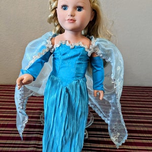 Puede incluir: Una mu&ntilde;eca princesa azul y plateada con cabello largo rubio y un vestido azul. La mu&ntilde;eca lleva una capa azul con ribetes plateados.