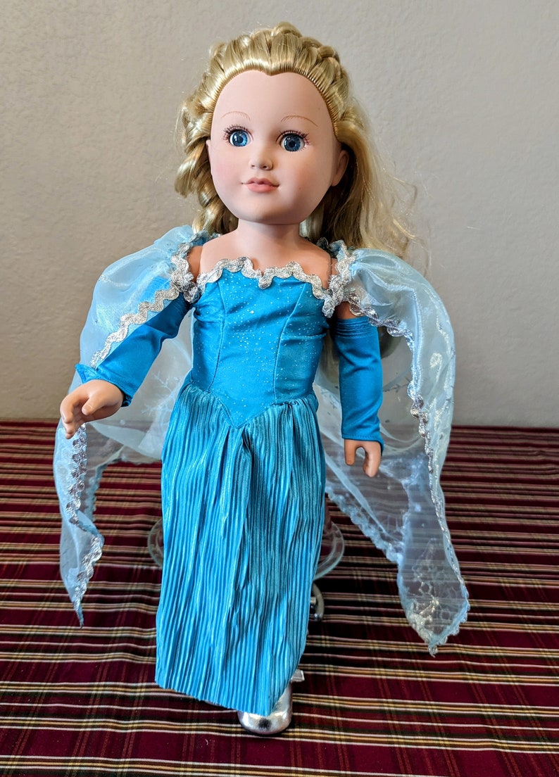 Puede incluir: Una mu&ntilde;eca princesa azul y plateada con cabello largo rubio y un vestido azul. La mu&ntilde;eca lleva una capa azul con ribetes plateados.