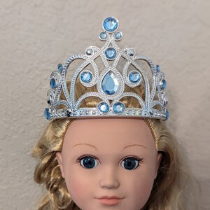 Puede incluir: Una tiara plateada con detalles de gemas azules. La tiara est&aacute; en la cabeza de una mu&ntilde;eca con cabello rubio.