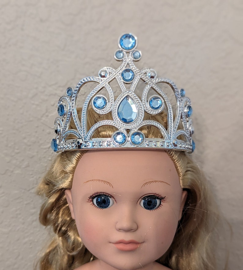 Puede incluir: Una tiara plateada con detalles de gemas azules. La tiara est&aacute; en la cabeza de una mu&ntilde;eca con cabello rubio.