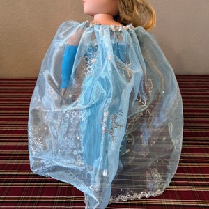 Puede incluir: Una capa de tul azul claro con detalles de copo de nieve plateados. La capa est&aacute; dise&ntilde;ada para una mu&ntilde;eca y tiene un borde festoneado.