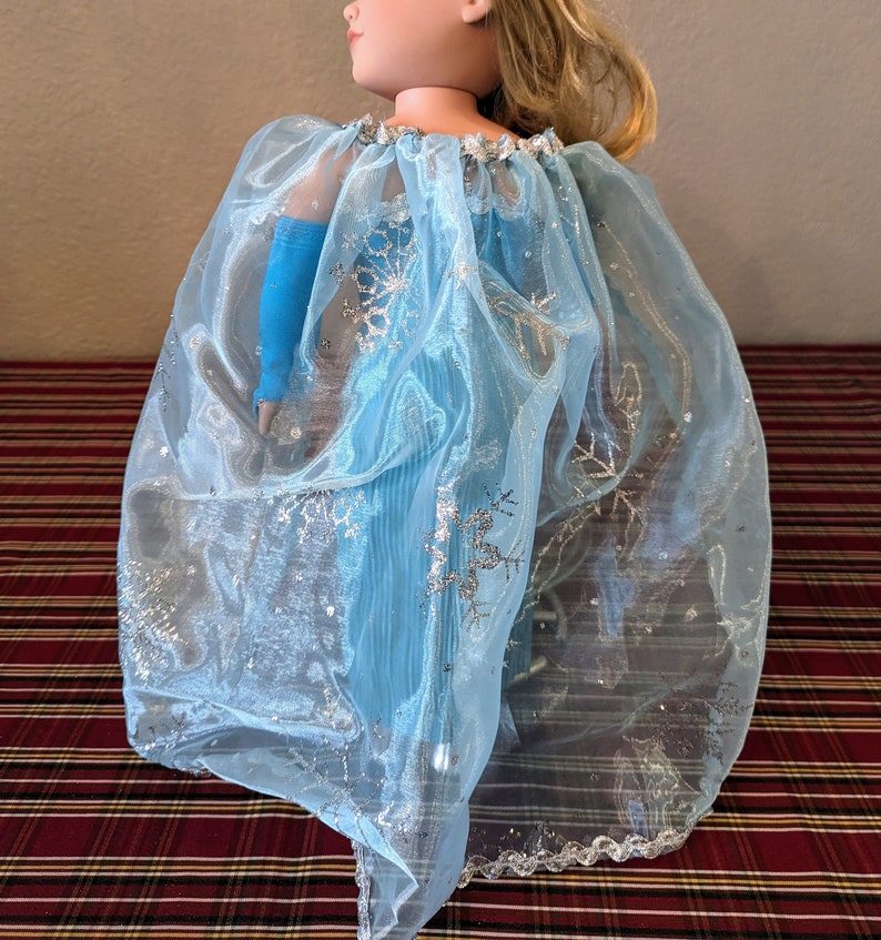 Puede incluir: Una capa de tul azul claro con detalles de copo de nieve plateados. La capa est&aacute; dise&ntilde;ada para una mu&ntilde;eca y tiene un borde festoneado.
