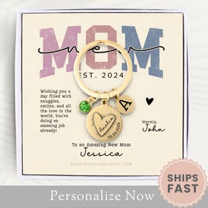 May include: A gold-toned keychain with a heart-shaped charm engraved with the name "Amelia" and the date "09.29.2021". The keychain also includes a small green gem and a letter "A" charm. The keychain is displayed in a white box with a pink and blue "MOM" design and the text "EST. 2024". The box also includes the text "Wishing you a day filled with snuggles, smiles, and all the love in the world. You're doing an amazing job already!" and "To an Amazing New Mom Jessica".