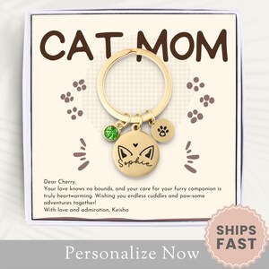 May include: A gold-toned keychain with a cat charm, a paw print charm, and a green gemstone. The keychain is in a white box with the words "CAT MOM" and a message for a cat owner. The box also has a "SHIPS FAST" sticker.