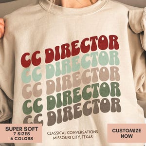 May include: A beige crewneck sweatshirt featuring the text CC DIRECTOR repeated five times in a stacked, wavy font. The text colors are red, light blue, tan, green, and brown. Below the text, the words CLASSICAL CONVERSATIONS MISSOURI CITY, TEXAS appear.