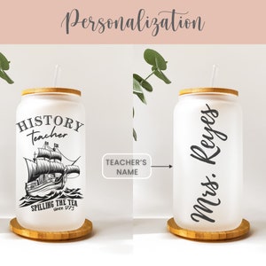 May include: A white frosted glass can with a bamboo lid and a straw. The can has a black and white illustration of a ship on the sea and the text "HISTORY teacher SPILLING THE TEA since 1773". The can on the right has the text "Mrs. Reyes" in black cursive.