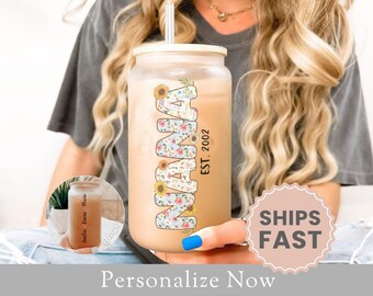 May include: A clear glass tumbler with a floral design and the word "MAMA" in colorful letters. The tumbler has a straw and a lid. The text "EST. 2002" is printed on the side of the tumbler.